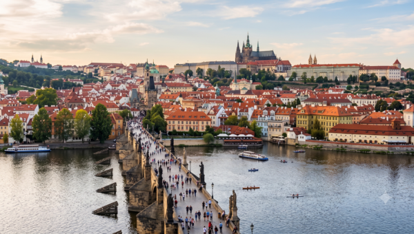 Panoramic view of Prague with Charles Bridge crowded with pedestrians, red-roofed houses, and Prague Castle on a hill above the Vltava River. (informative)